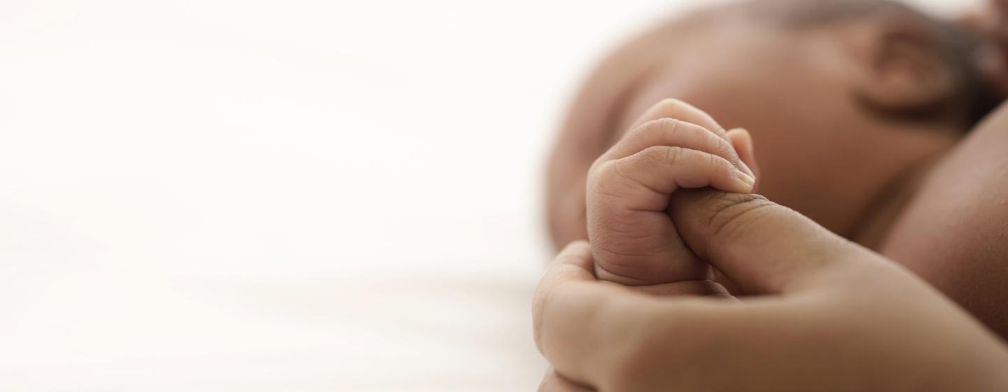 A Black infant is lying on the bed while its mother holds its hand.